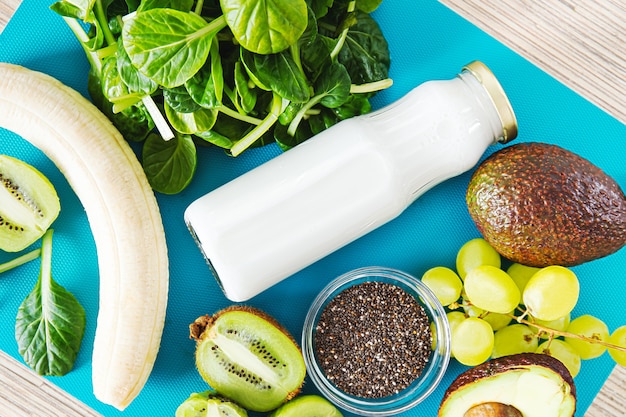 Flat lay ingredients for a green smoothie. Top view on a turquoise background of banana, spinach, chia seeds, coconut milk in a glass jar, avocado, grapes. Detox, smoothie recipe for a healthy diet.