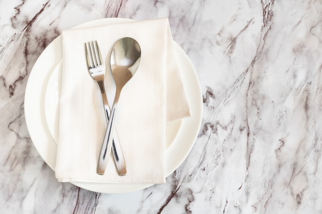 Flat lay is cutlery, fork and knife on a napkin on an empty white plate on a marble table.