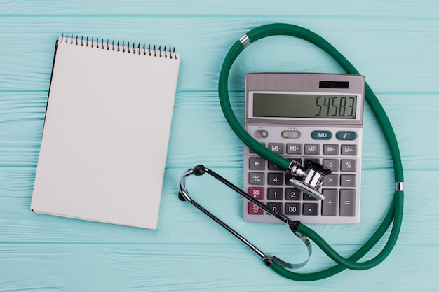 Flat lay medical composition include calculator notepad stethoscope on a blue pastel background. White blank notepad. From top view.