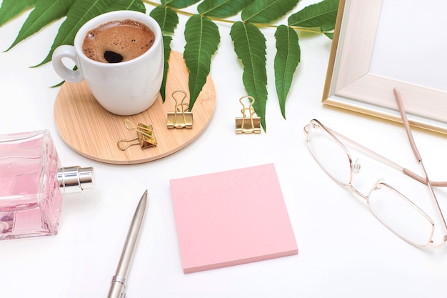 Flat lay, office desk, top view. Workspace with a coffee mug, an angry plant leaf, a perfume bottle, gold glasses and a pen, and a pink sticker for text on a white background. Woman home office