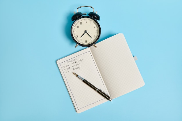 Flat lay of an open organizer with plans for the day, ink pen and alarm clock on blue background with copy space. Time management, business and planning concept