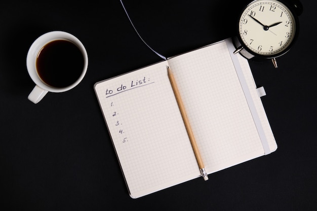 Flat lay of an opened organizer, notepad with list to do on blank white sheets, cup of coffee and alarm clock on black background with copy space. Business, Organization, Time Management concepts..