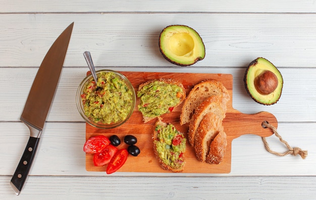 Flat lay photo, freshly prepared guacamole in small glass bowl, bread, tomatoes, olives at working board and two avocados next white wood desk