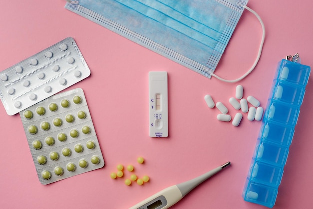 Flat lay of pills covid19 test medical mask and thermometer on pink background