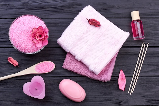 Flat lay pink bath body care accessories on dark wood. Salt with towel and soap.