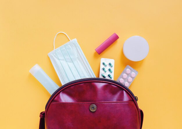 Flat lay of red leather woman bag open out with cosmetics, accessories and mask with alcohol gel sanitizer on yellow