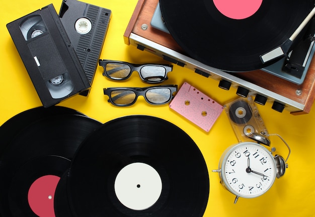 Flat lay retro style attributes, 80s media. Vinyl player, video cassettes, audio cassettes, records, 3d glasses, vintage alarm clock, old books on yellow background. Top view