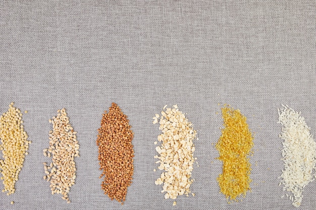 Photo flat lay of set of heap various grains and cereals bulgur, rice, oatmeal, buckwheat, couscous wheat seeds on grey background, copy space, space for text, top view.