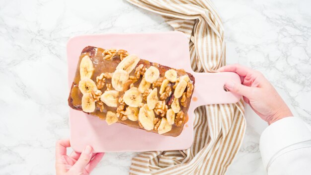 Flat lay. Slicing banana nut bread topped with caramel and garnished with banana chips and walnuts.