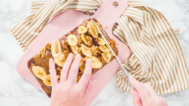 Flat lay. Slicing banana nut bread topped with caramel and garnished with banana chips and walnuts.