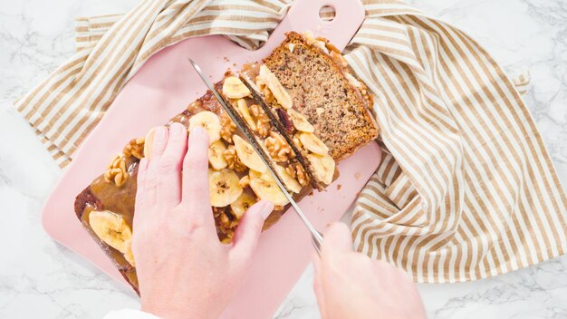 Flat lay. Slicing banana nut bread topped with caramel and garnished with banana chips and walnuts.