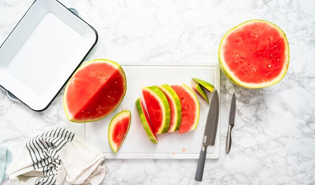 Flat lay. Slicing red seedless watermelon on a white cutting board.