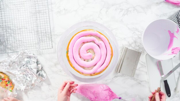 Flat lay. Step by step. Frosting round funfetti cake layers with pink buttercream frosting.