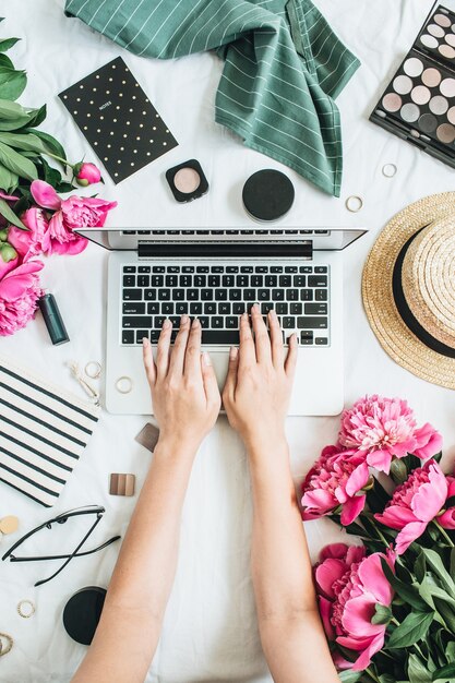 Flat lay styled office desk with laptop, pink peony flowers, cosmetics, accessories