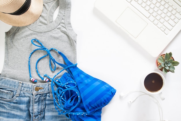 Flat lay of summertime clothing with blue stripe bikini, laptop and cup of coffee on white background