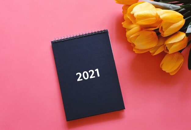 Flat lay top view of black diary or planner 2021 with yellow tulip flower on red background with copy space, new year resolutions concept