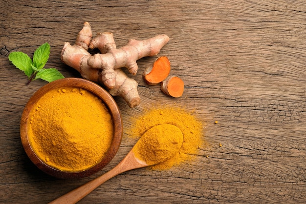 Flat lay (top view) of turmeric (curcumin) powder in wooden bowl and spoon with fresh rhizome on wood background.