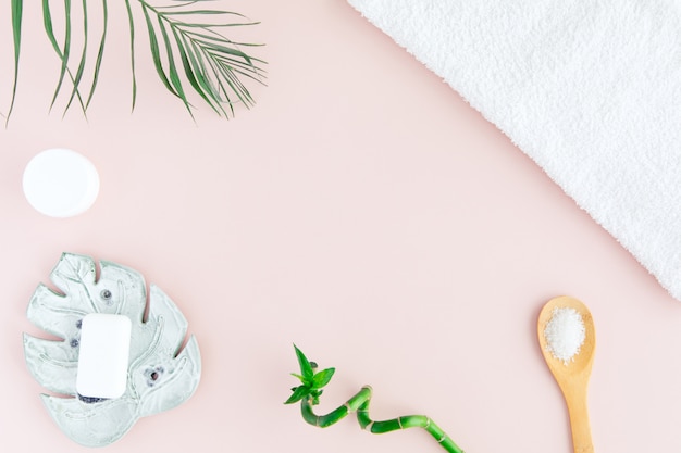 Flat lay and top view of white towel, jar of cream, green palm leaves and bamboo on pastel pink