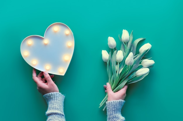 Flat lay, top view with hand showing plastic heart shaped lightboard with lights and bunch of white tulips