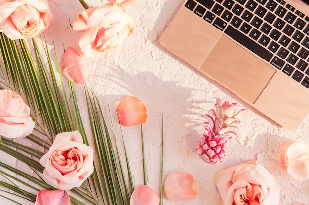 Flat lay of tropical workspace mockup with modern laptop, palm monstera leaves, pink flowers, exotic pineapple and petals on pastel