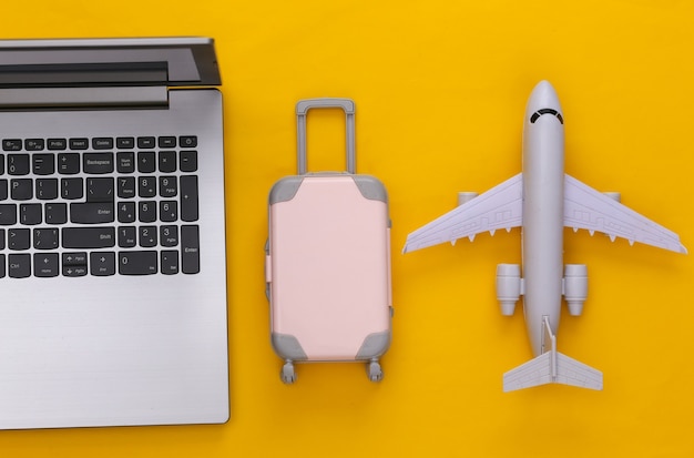 Flat lay vacation holiday and travel planing concept. Laptop and mini plastic travel suitcase, air plane on yellow background. Top view