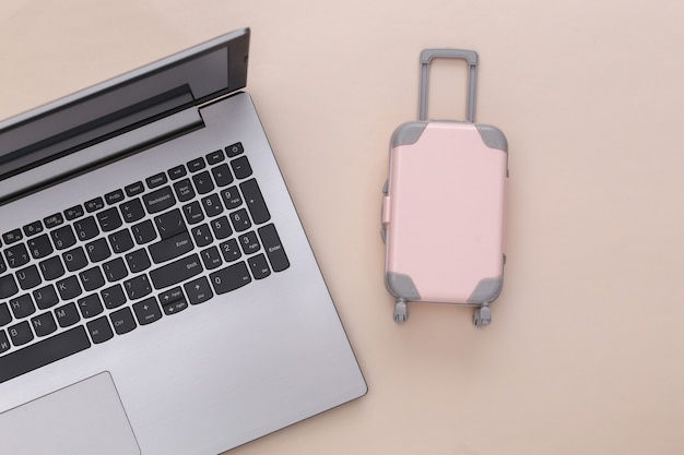 Flat lay vacation holiday and travel planing concept. Laptop and mini plastic travel suitcase on beige background. Top view