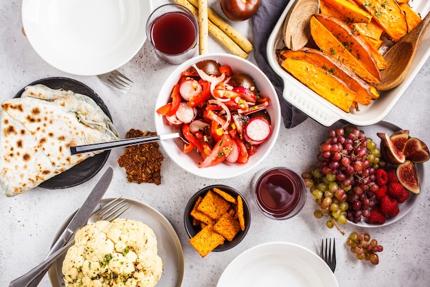 Piatto lay di cibo vegano: patate dolci al forno, cavolfiore, frutta, insalata di verdure e tortilla con verdure su sfondo bianco.