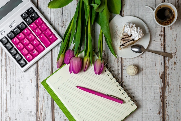 Flat lay view of the surface of the desktop holiday in the\
financial sphere tulips coffee with a cake and a notepad for notes\
with a calculator on a wooden light background