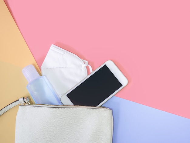 Flat lay of white leather woman bag with smartphone, mask and alcohol gel sanitizer