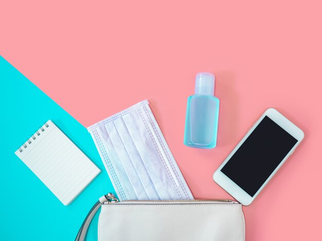 Disposizione piana della borsa della donna del cuoio bianco con il disinfettante del gel dello smartphone, della maschera chirurgica, del taccuino e dell'alcool su fondo variopinto, vista superiore con lo spazio della copia per testo. concetto di covid-19 e coronavirus.