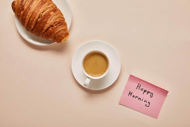 Flat lay with coffee croissant and sticky note with happy morning lettering on beige background