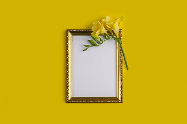 Flat lay with an empty golden photo frame and a delicate freesia flower on a yellow background