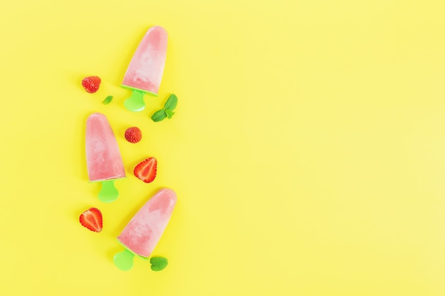 Piatto disteso con gelato sorbetto congelato rosa fruttato su raccolta di bastoncini di plastica verde con tagli di fragola, foglie di menta su sfondo giallo. frutta mista surgelata con yogurt sotto forma di gelato