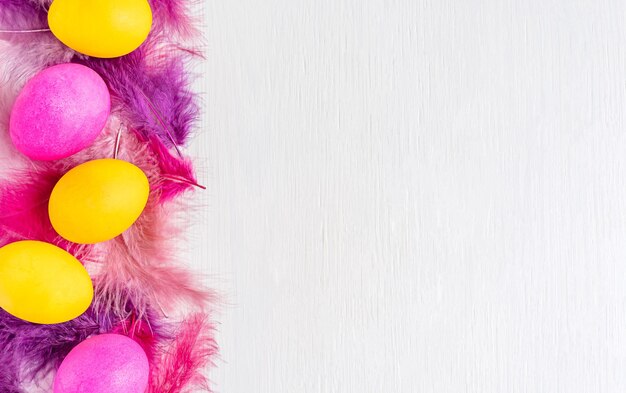 Фото Плоская лежала с окрашенными в ярко-розовые и желтые пасхальные яйца с фиолетовыми перьями на белом деревянном столе