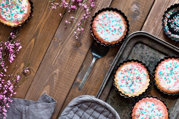 Flat lay with small parrotot cakes with sprinkled sugar on top with small pink flowers on metal baking tray and dark wood