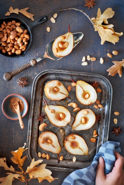 Flat lay with a tray of baked pears with caramelized nuts on dark wooden background with Autumn leaves