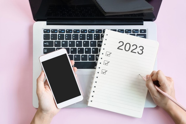 Photo flat lay of woman hand writing 2023 to do list on notebook while holding mobile phone with blank screen copy space top view 2023 planning concept