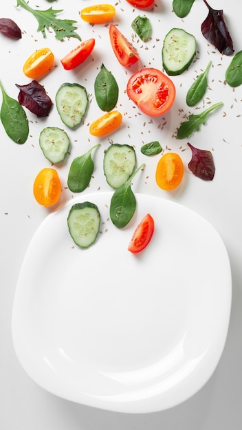 Flat layered fresh vegetables, herbs and spices with an empty white plate on a white background.