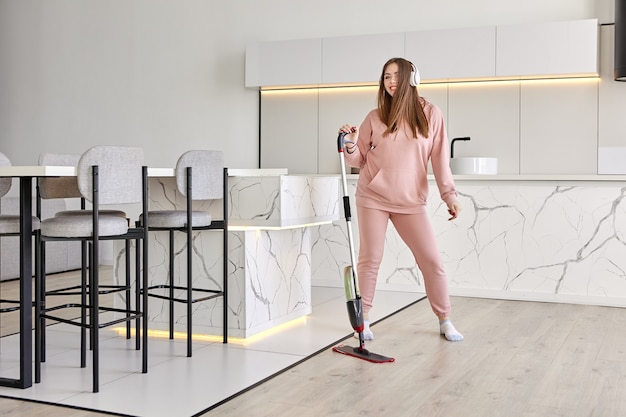 Flat mop with spray and microfiber pad in hands of young woman who is listening to music, tidying up and dancing in kitchen.