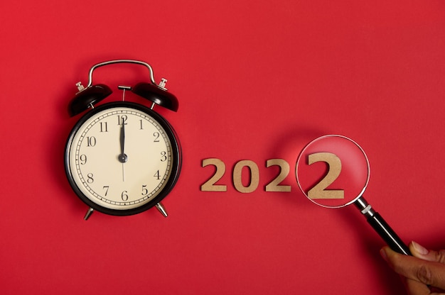 A flat view of midnight on a black alarm clock face next to wooden numerals and a cropped hand holding a magnifying glass showing the year 2022. New year concept isolated over red background