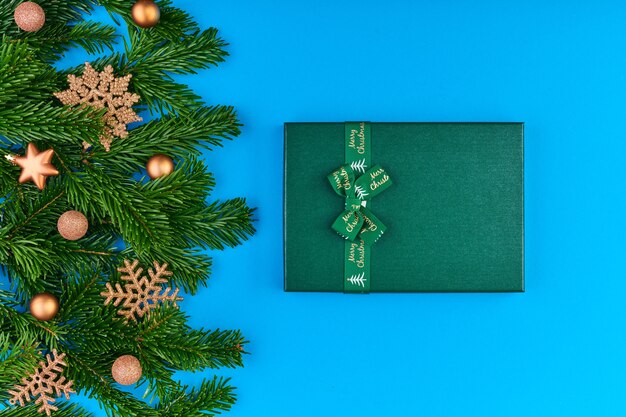 Flatlay of golden decorative snowflakes, stars and balls on evergreen coniferous branches and giftbox with xmas present on blue background