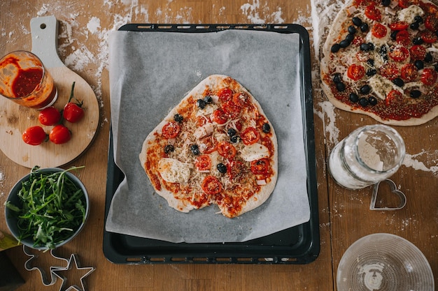 Photo flatlay of heart shaped pizza dough covered with pizza sauce and ingredients on wooden table