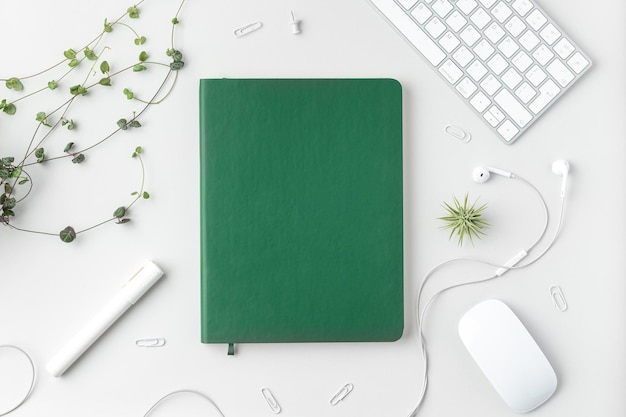 Photo flatlay of home office desk table top view of workspace with green notebook keyboard mouse marker headphones pins and plants on white background