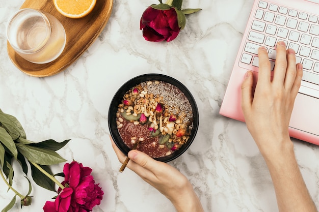 Flatlay женских рук, держа чашу смузи с ноутбуком, пионов и воды на мраморном столе