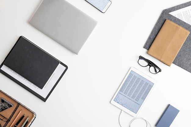 Flatlay of tablet, documents in clipboard, several notebooks in leather cover, eyeglasses, smartphone, envelope with paper and office supplies