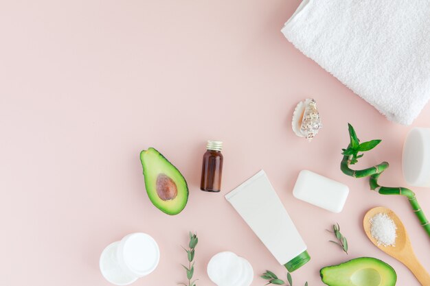 Flatlay and top view of avocado, avocado oil, white towel, jar of cream, green leaves and bamboom avocado on pastel pink