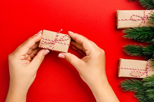 Flatlay van vrouwelijke handen met geschenkdoos gemaakt van ambachtelijk papier op rode tafel met kerstversieringen