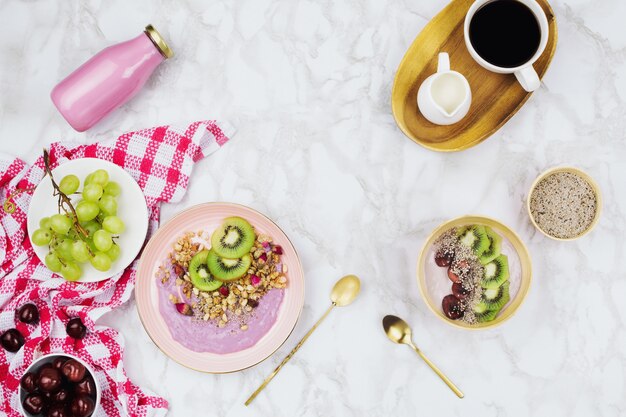 Flatlay of vegan breakfast with plant based yogurt bowls topped with kiwi slices, granola, chia seeds, smoothie bottle and coffee with soy milk on marble background