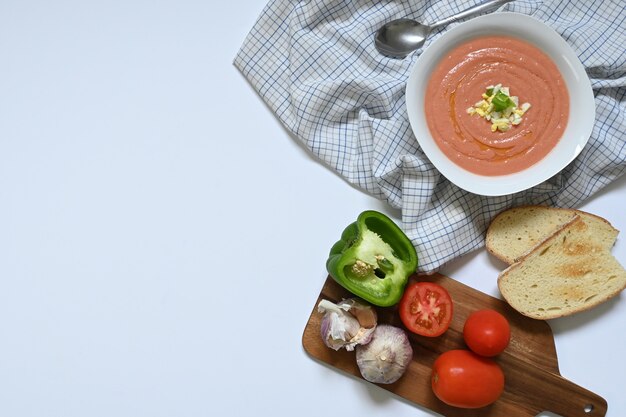 Flatlay of white plate with Spanish cordovan salmorejo gazpacho next to dark wood table with tomato, green pepper, garlic and toast, on a checkered tablecloth and a white surface, with copy space