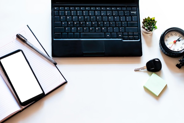 Flatlay della scrivania dell'area di lavoro bianca con laptop nero, blocco note, schermo bianco del telefono cellulare, orologio, prodotti lattiero-caseari